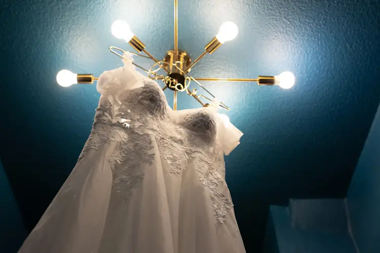 Ein weißes Spitzenbrautkleid hängt von unten betrachtet auf einem Bügel, beleuchtet von einem modernen goldenen Kronleuchter mit freiliegenden Glühbirnen vor einer türkisfarbenen Decke. Das Kleid, wunderschön eingefangen vom Hochzeitsfotografen Dresden, besticht durch aufwendige florale Spitzendetails.