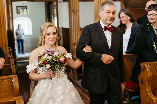 Eine Braut in einem weißen Brautkleid mit einem Blumenstrauß in der Hand geht mit einem älteren Mann im Anzug den Gang entlang. Sie befinden sich in einer Kirche, umgeben von sitzenden Gästen.