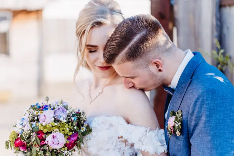 Ein Bräutigam im blauen Anzug küsst sanft die Schulter der Braut, während sie einen bunten Blumenstrauß hält und lächelt. Beide stehen an ihrem Hochzeitstag im Freien eng beieinander.