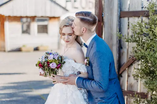 Ein Bräutigam im blauen Anzug küsst seine Braut auf die Stirn. Sie hält einen bunten Blumenstrauß in der Hand und trägt ein weißes, schulterfreies Brautkleid. Sie stehen im Freien in der Nähe eines rustikalen Holzgebäudes und eines Tors.