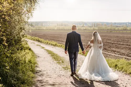 Braut und Bräutigam gehen Hand in Hand einen Feldweg entlang, der an einem Feld entlangführt. Auf der einen Seite ist Grün, und das Sonnenlicht scheint auf ihre Rücken. Die Braut trägt ein weißes Kleid und einen Schleier; der Bräutigam trägt einen dunklen Anzug.