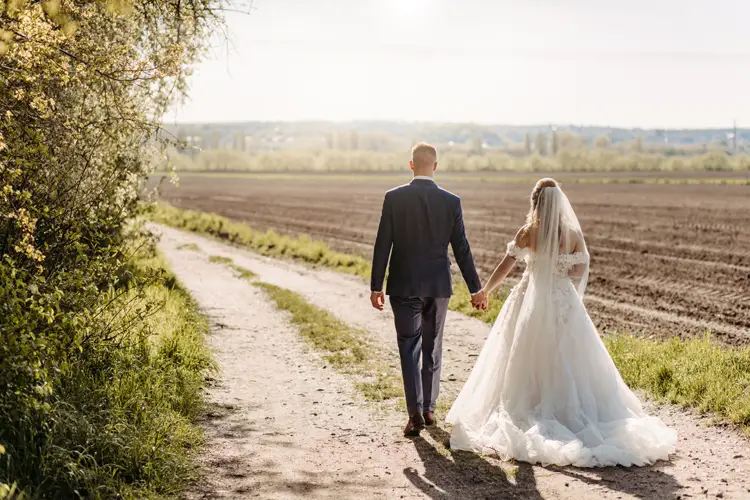 Braut und Bräutigam gehen Hand in Hand einen Feldweg entlang, der an einem Feld entlangführt. Auf der einen Seite ist Grün, und das Sonnenlicht scheint auf ihre Rücken. Die Braut trägt ein weißes Kleid und einen Schleier; der Bräutigam trägt einen dunklen Anzug.