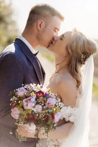 Braut und Bräutigam küssen sich liebevoll im Freien. Die Braut hält einen Strauß rosa und lila Blumen in der Hand und trägt ein weißes Kleid mit Schleier, während der Bräutigam einen dunklen Anzug trägt. Das Sonnenlicht erhellt das Paar sanft.