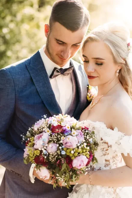 Braut und Bräutigam stehen im Freien eng beieinander. Beide lächeln sanft und halten einen Strauß rosa, violetter und weißer Blumen in den Händen. Das Sonnenlicht erhellt sanft ihre Gesichter und schafft eine romantische Atmosphäre.