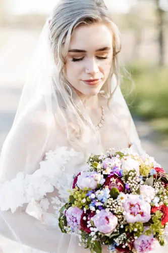 Eine Braut mit langem blondem Haar und einem transparenten Schleier blickt nach unten, während sie im Freien im sanften Sonnenlicht einen Strauß rosa, violetter und weißer Blumen hält.