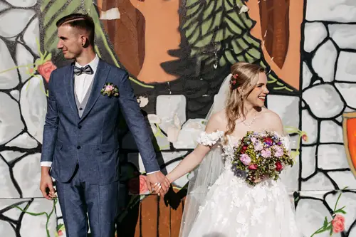 Eine Braut im weißen Kleid hält einen bunten Blumenstrauß in der Hand, und ein Bräutigam im blauen Anzug hält sich an den Händen, während sie vor einer farbenfrohen Wand im Graffiti-Stil stehen. Beide blicken in entgegengesetzte Richtungen.