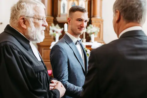 Ein Bräutigam in einem blauen Anzug steht zwischen zwei älteren Männern in Anzügen in einer Kirche, lächelt und unterhält sich vor einem mit Blumen geschmückten Holzalt.