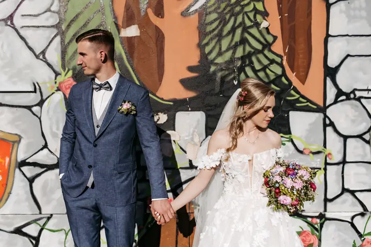 Eine Braut im weißen Kleid hält einen bunten Blumenstrauß in der Hand, und ein Bräutigam im blauen Anzug hält sich an den Händen, während sie vor einer farbenfrohen Wand im Graffiti-Stil stehen. Beide blicken in entgegengesetzte Richtungen.