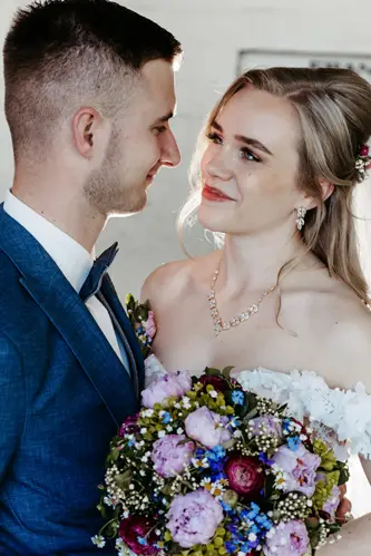 Braut und Bräutigam blicken sich liebevoll an. Die Braut trägt ein schulterfreies weißes Kleid, Schmuck und einen bunten Blumenstrauß in der Hand. Der Bräutigam trägt einen blauen Anzug mit Fliege. Beide lächeln in einem hellen Innenraum.