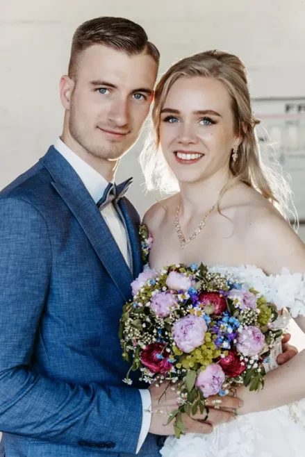 Braut und Bräutigam stehen dicht beieinander und lächeln in die Kamera. Die Braut hält einen bunten Blumenstrauß in der Hand und trägt ein weißes Kleid, während der Bräutigam einen blauen Anzug mit Fliege trägt.