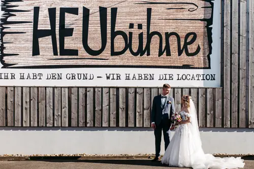 Braut und Bräutigam stehen vor einem großen Holzschild mit der Aufschrift „HEUbühne“ und deutschem Text darunter. Die Braut hält einen bunten Blumenstrauß in der Hand und trägt ein weißes Kleid mit Schleier, während der Bräutigam einen Anzug trägt.