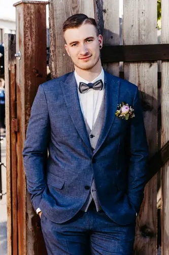 Ein Mann im blauen Anzug mit Fliege und Ansteckblume steht im Freien, lehnt an einem Holzzaun und blickt mit einem leichten Lächeln in die Kamera.