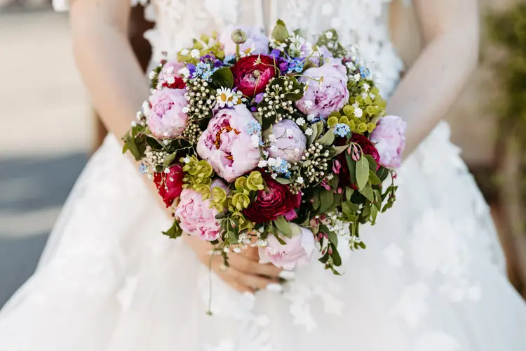Eine Braut in einem weißen Spitzenkleid hält einen farbenfrohen Strauß aus rosa Pfingstrosen, roten und weißen Blumen, blauen Akzenten und Grünpflanzen.