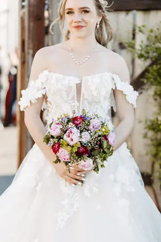 Eine Braut in einem schulterfreien weißen Spitzenbrautkleid hält einen bunten Strauß aus rosa, roten und violetten Blumen und lächelt sanft im Freien.