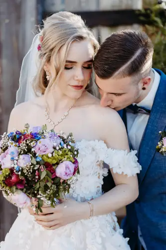 Eine Braut in einem schulterfreien weißen Kleid hält einen bunten Blumenstrauß und blickt sanft lächelnd nach unten, während ein Bräutigam in einem dunklen Anzug seinen Kopf sanft zu ihrer Schulter neigt.