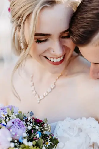 Eine Braut in einem schulterfreien weißen Kleid hält einen bunten Blumenstrauß und blickt sanft lächelnd nach unten, während ein Bräutigam in einem dunklen Anzug seinen Kopf sanft zu ihrer Schulter neigt.
