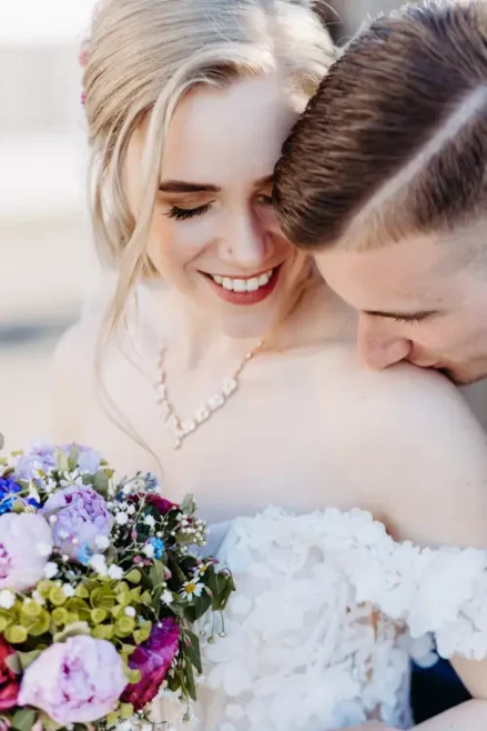 Eine Braut in einem schulterfreien weißen Kleid hält einen bunten Blumenstrauß und blickt sanft lächelnd nach unten, während ein Bräutigam in einem dunklen Anzug seinen Kopf sanft zu ihrer Schulter neigt.