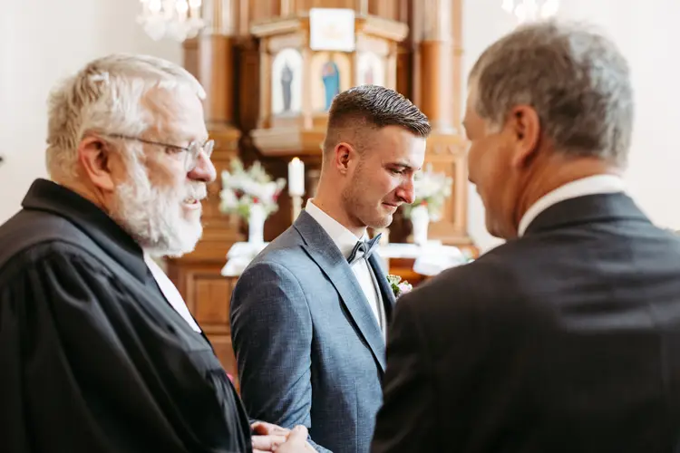Ein Bräutigam in einem blauen Anzug steht zwischen zwei älteren Männern in Anzügen in einer Kirche, lächelt und unterhält sich vor einem mit Blumen geschmückten Holzalt.