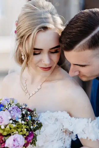 Eine Braut in einem schulterfreien weißen Kleid hält einen bunten Blumenstrauß und blickt sanft lächelnd nach unten, während ein Bräutigam in einem dunklen Anzug seinen Kopf sanft zu ihrer Schulter neigt.