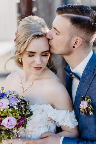 Eine Braut in einem schulterfreien weißen Kleid mit einem bunten Blumenstrauß steht eng neben ihrem Bräutigam im blauen Anzug. Beide haben die Augen geschlossen und lächeln sanft. Sie wirken entspannt und glücklich.