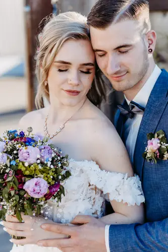 Eine Braut in einem schulterfreien weißen Kleid mit einem bunten Blumenstrauß steht eng neben ihrem Bräutigam im blauen Anzug. Beide haben die Augen geschlossen und lächeln sanft. Sie wirken entspannt und glücklich.
