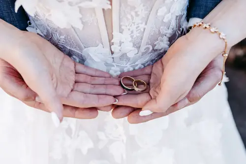 Eine Nahaufnahme von zwei Händen, die sanft ein Paar goldene Eheringe halten, mit einem weißen Spitzenbrautkleid und einem zarten Armband im Hintergrund.