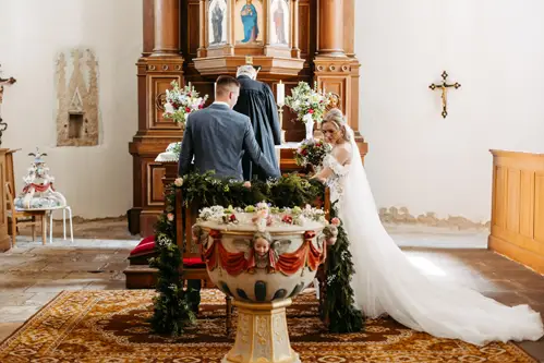 Braut und Bräutigam stehen in einer Kirche am Altar und blicken einem Priester entgegen. Die Braut trägt ein weißes Kleid mit langer Schleppe, und der Altar ist mit Blumen und Grünpflanzen geschmückt. Im Vordergrund steht ein kunstvolles Taufbecken.