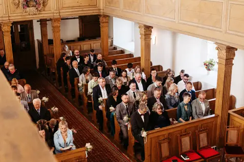 Eine Gruppe formell gekleideter Menschen sitzt und steht in Holzbänken in einer Kirche und nimmt offenbar an einer Hochzeitszeremonie teil. Einige halten kleine Blumenarrangements in den Händen, und Licht fällt durch die Fenster.