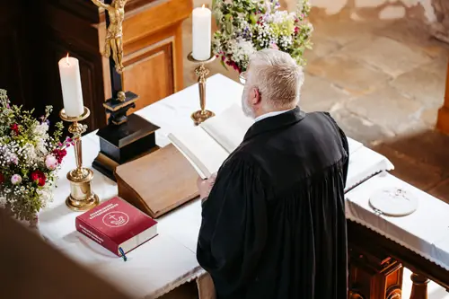 Ein Geistlicher in einer schwarzen Robe steht mit Kerzen, Blumen, einem offenen Buch und einem roten Gesangbuch an einem Altar und bereitet wahrscheinlich einen Gottesdienst in einer Kirche vor oder leitet ihn.