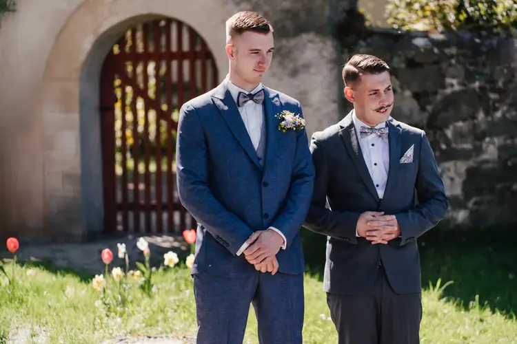 Zwei Männer in Anzügen stehen draußen in der Nähe einer Steinmauer und eines Torbogens. Im Hintergrund sind Blumen und Grünpflanzen zu sehen. Ein Mann trägt einen blauen Anzug mit Ansteckblume, der andere einen dunklen Anzug mit gemusterter Fliege.