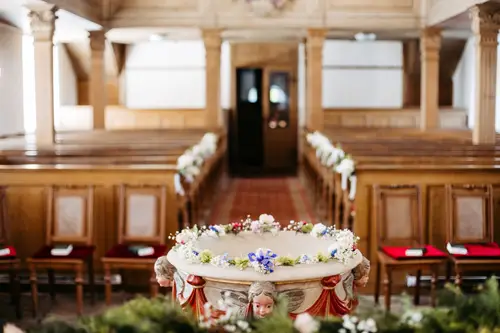 Ein mit Blumen geschmücktes Taufbecken steht vor dem leeren, hölzernen Kirchenraum, der mit Kirchenbänken gesäumt ist. Natürliches Licht strömt durch die Fenster und unterstreicht die friedliche, zeremonielle Atmosphäre.