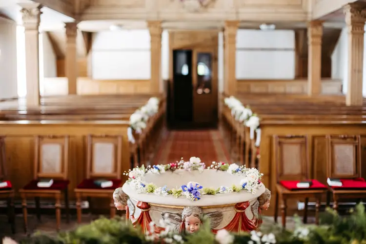 Ein mit Blumen geschmücktes Taufbecken steht vor dem leeren, hölzernen Kirchenraum, der mit Kirchenbänken gesäumt ist. Natürliches Licht strömt durch die Fenster und unterstreicht die friedliche, zeremonielle Atmosphäre.