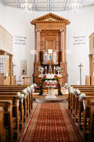 Ein dekorierter Kirchenraum mit Holzbänken, einem rot gemusterten Gangläufer und einem großen Holzaltar, geschmückt mit Blumen, Kerzen und religiösen Ikonen. Die weißen Wände weisen beidseitig deutsche Inschriften auf.