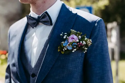 Ein Mann in blauem Anzug und Fliege trägt eine große, farbenfrohe Blumenansteckblume am Revers seines Jacketts. Der Hintergrund ist im Freien und leicht unscharf.