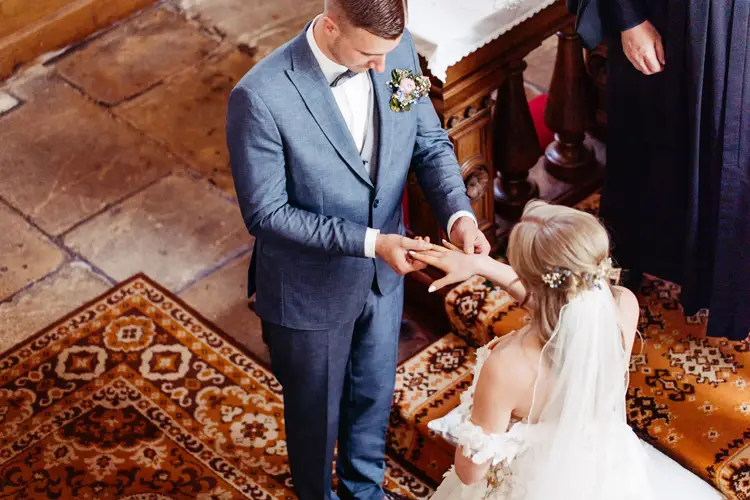 Ein Bräutigam im blauen Anzug steckt der Braut während einer Hochzeitszeremonie einen Ring an den Finger (von oben gesehen). Die Braut trägt ein weißes Kleid mit Schleier und sie stehen auf einem gemusterten Teppich.