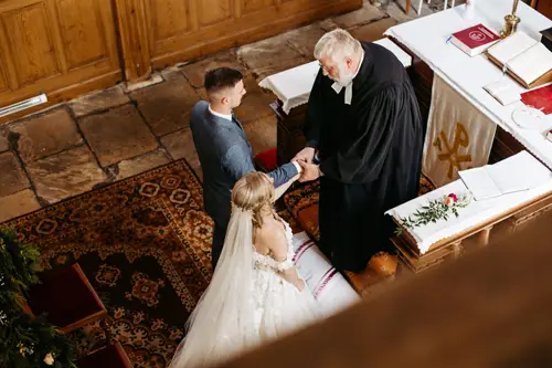 Braut und Bräutigam stehen während ihrer Hochzeitszeremonie Hand in Hand vor einem Pfarrer in einer Kirche (von oben gesehen). Der Altar ist mit Büchern, einem Tuch und einer einzelnen Rose geschmückt.