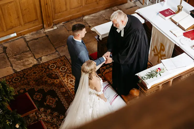 Braut und Bräutigam stehen während ihrer Hochzeitszeremonie Hand in Hand vor einem Pfarrer in einer Kirche (von oben gesehen). Der Altar ist mit Büchern, einem Tuch und einer einzelnen Rose geschmückt.