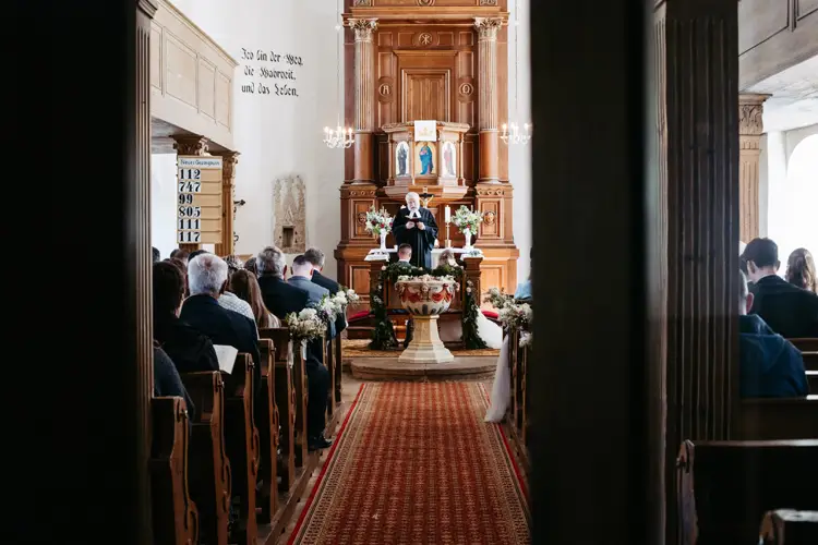 Eine Gemeinde sitzt in Holzbänken gegenüber einem Pfarrer, der am Altar einer traditionellen Kirche mit hohen Holzpaneelen und Blumenschmuck spricht. Licht strömt durch die Fenster und erhellt den Innenraum.