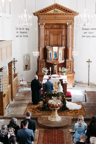 Eine Hochzeitszeremonie findet in einer Kirche statt. Braut und Bräutigam stehen neben einem Priester vor dem Altar. Die Gäste nehmen Platz, und der Kirchenraum ist mit Kronleuchtern, Holzelementen und religiösem Dekor geschmückt.