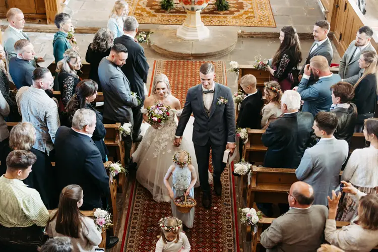 Ein Brautpaar schreitet in einer Kirche händchenhaltend und lächelnd den Gang entlang, umgeben von sitzenden Gästen. Blumenmädchen mit Blumenkränzen und Blumenkörben führen den Weg. Die Kirchenbänke sind mit Blumen geschmückt.