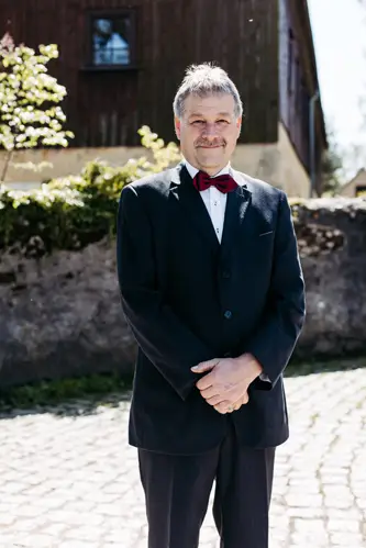 Ein Mann mittleren Alters mit kurzen grauen Haaren, der einen schwarzen Anzug, ein weißes Hemd und eine rote Fliege trägt, steht an einem sonnigen Tag im Freien vor einer Steinmauer und einem Holzgebäude.