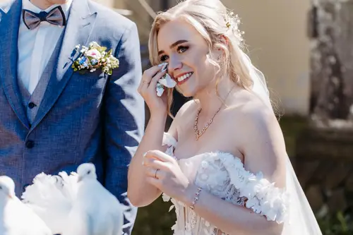 Eine Braut in einem weißen, schulterfreien Brautkleid lächelt und wischt sich eine Träne aus dem Gesicht. Sie steht neben einem Bräutigam im blauen Anzug mit einer Blumenboutonniere. Im Vordergrund sind weiße Tauben zu sehen.