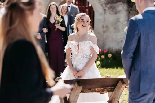 Eine Braut in einem weißen schulterfreien Kleid lächelt ihren Bräutigam während einer Hochzeitszeremonie im Freien an, während die Gäste im Hintergrund stehen und zuschauen.