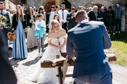 Eine Braut in einem weißen Kleid und ein Bräutigam in einem blauen Anzug verwenden eine Säge, um gemeinsam einen Baumstamm im Freien zu zersägen, umgeben von lächelnden Hochzeitsgästen an einem sonnigen Tag.