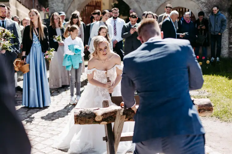 Eine Braut in einem weißen Kleid und ein Bräutigam in einem blauen Anzug verwenden eine Säge, um gemeinsam einen Baumstamm im Freien zu zersägen, umgeben von lächelnden Hochzeitsgästen an einem sonnigen Tag.