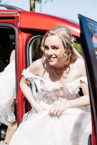 Eine lächelnde Braut in einem weißen schulterfreien Brautkleid steigt an einem sonnigen Tag aus einem roten Auto, hält ihr Kleid in der Hand und blickt aufgeregt nach vorne.