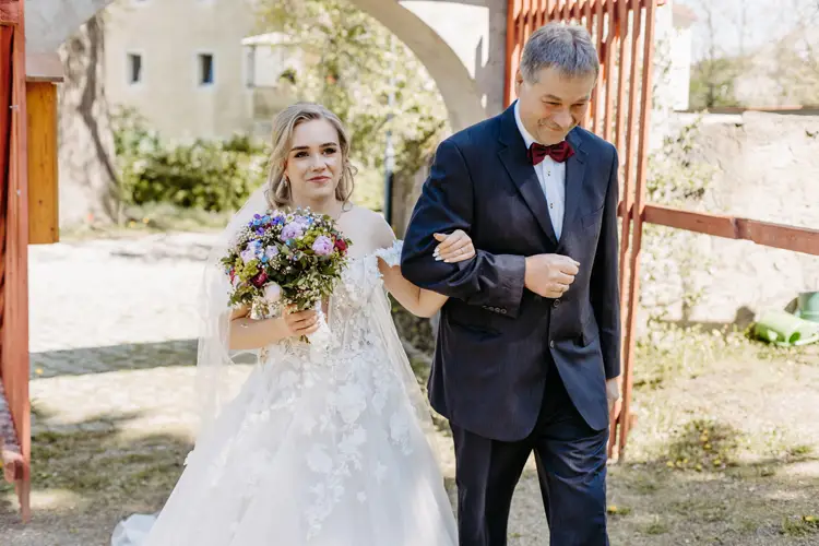 Eine lächelnde Braut in einem weißen, schulterfreien Kleid hält einen bunten Blumenstrauß und geht Arm in Arm mit einem älteren Mann in einem dunklen Anzug und einer roten Fliege im Freien in einem Garten spazieren.
