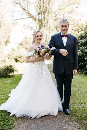 Eine lächelnde Braut in einem weißen, schulterfreien Kleid hält einen bunten Blumenstrauß und geht Arm in Arm mit einem älteren Mann in einem dunklen Anzug und einer roten Fliege im Freien in einem Garten spazieren.