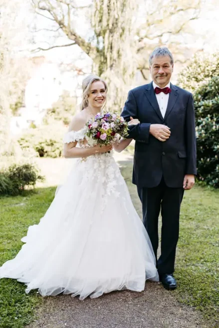 Eine lächelnde Braut in einem weißen, schulterfreien Kleid hält einen bunten Blumenstrauß und geht Arm in Arm mit einem älteren Mann in einem dunklen Anzug und einer roten Fliege im Freien in einem Garten spazieren.