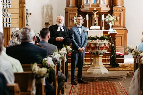 Ein Bräutigam im hellblauen Anzug steht neben einem Pfarrer in einer Kirche und blickt auf sitzende Gäste. Der Kirchenraum ist mit Blumen und Holzmöbeln geschmückt, im Vordergrund steht ein kunstvolles Taufbecken.